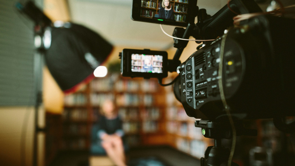 Gros plan sur une caméra de tournage et son moniteur, un interviewé assis flou dans une bibliothèque
