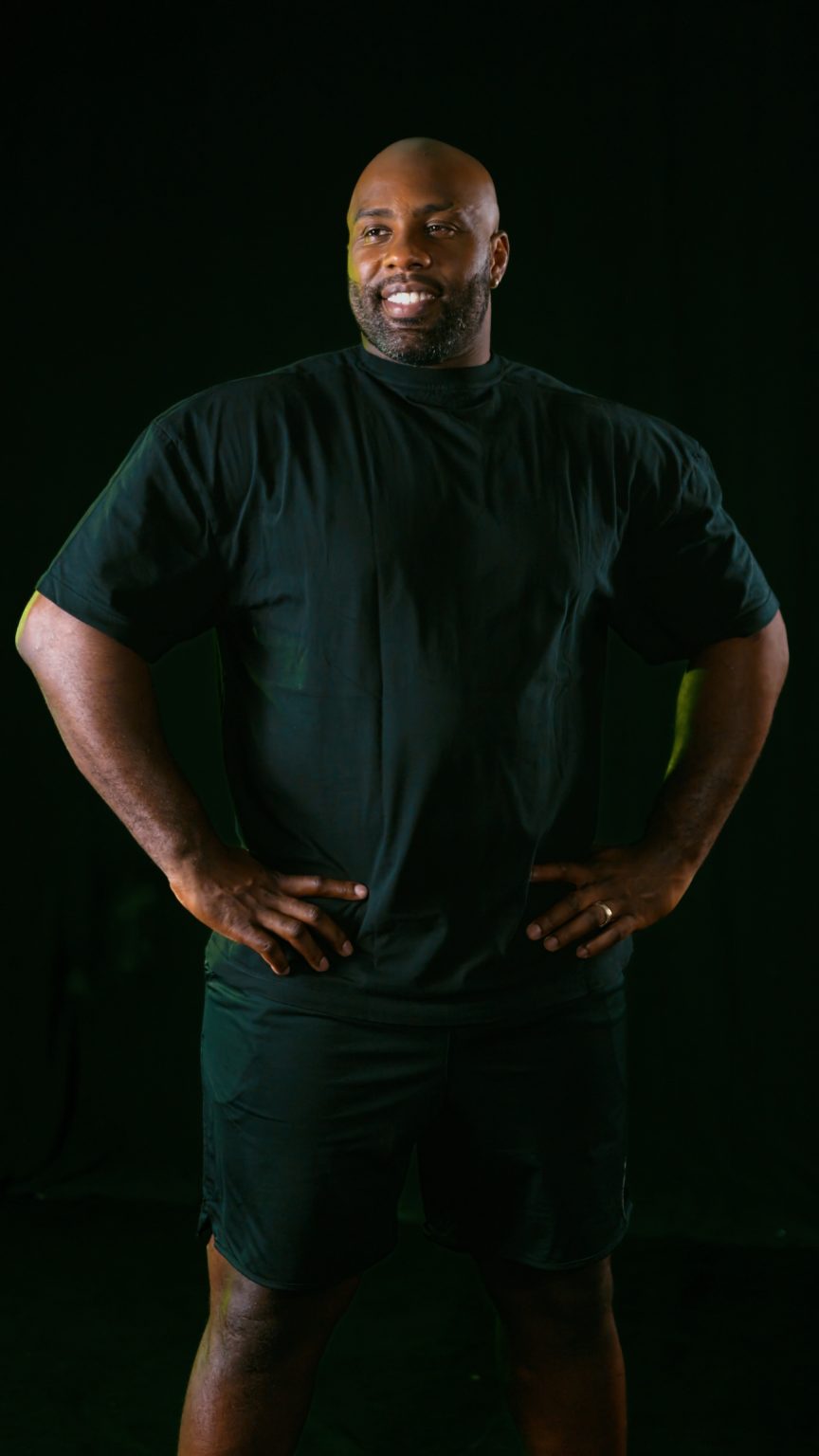 Teddy Riner smiling, hands on hips, against a dark background lit in green
