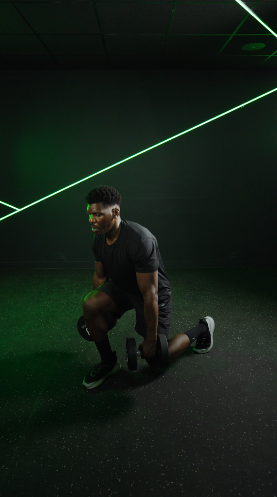 An athlete in a kneeling lunge, dumbbells in hand, under a green neon light
