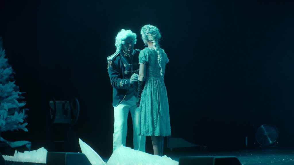 Two costumed actors, one in white and the other in a blue gown, facing each other in blue light