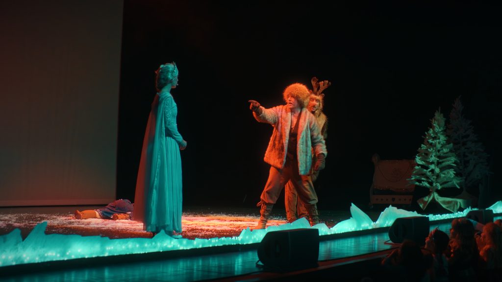An actor in a reindeer costume and an actress in a turquoise gown on stage, under orange light