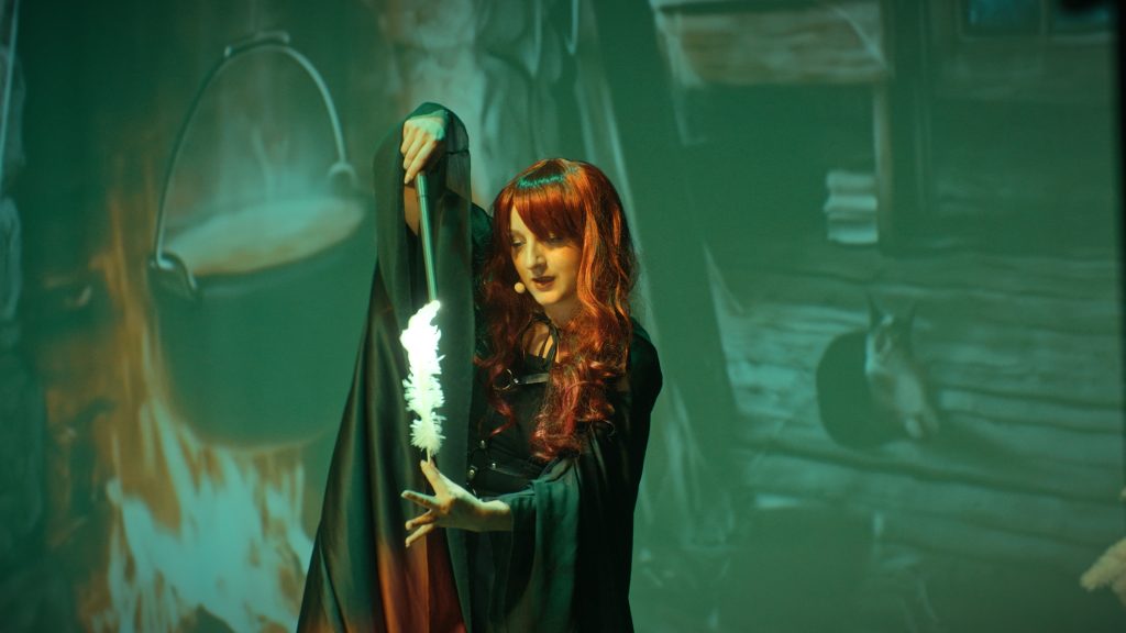 A red-haired actress in a dark outfit in front of a flaming cauldron