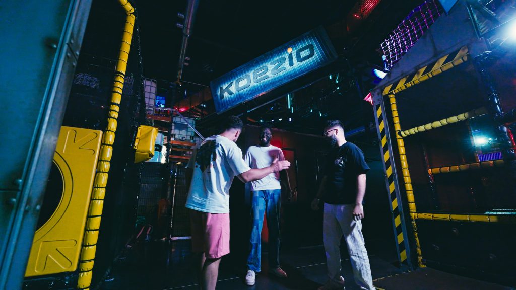 Three visitors at the entrance of a game room with metal structures and the Koezio sign