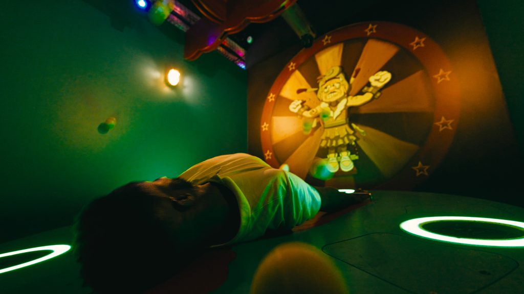 A player lying on the floor facing an illustrated light wheel, in a room lit in green