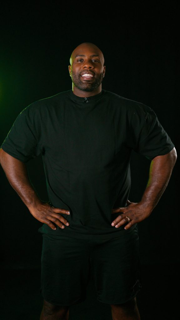 Teddy Riner smiling, hands on hips, against a dark background lit in green